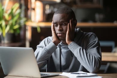 tired african american man working on laptop in coffeehouse, holding head in hands, looking at screen, exhausted student taking break, watching webinar, working on research work or homework in cafe