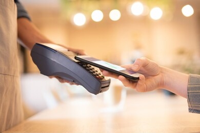 young businesswoman holding her smartphone close to keypad of electronic payment machine held by waiter of cafe