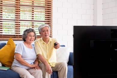 asian couple senior sitting on sofa and use remote control to change channel and watching tv in living room at home.panning from blur foreground television