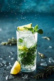 fresh mojito cocktail with lime, rosemary, mint and ice in jar glass on dark blue background. studio shot of drink in freeze motion, drops in liquid splash. summer cold drink and cocktail