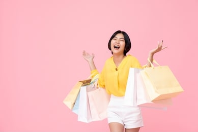 happy attractive young thai asian female carrying pastel colored shopping bags with both arms raised in a ecstatic gesture isolated in pink studio background