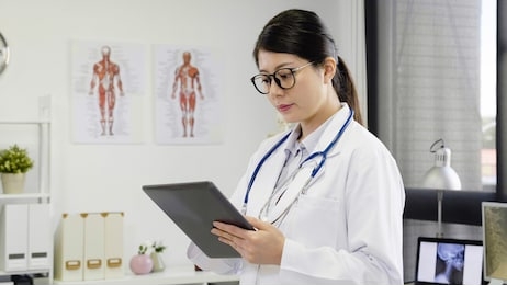 asian woman doctor in glasses using tablet computer while standing straight in hospital. young female medical staff hands finger touching on digital pad screen concentrated working in clinic office.