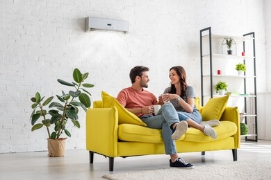 happy young couple talking and holding cups while sitting on yellow sofa under air conditioner at home