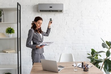 beautiful businesswoman reading business newspaper while standing under air conditioner with remote control
