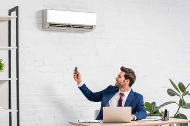 handsome businessman holding remote control while sitting at workplace under air conditioner