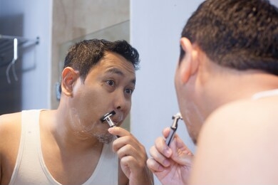 portrait of attractive young asian man shaving in bathroom, smiling and looking at mirror