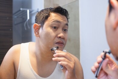 portrait of attractive young asian man shaving in bathroom, smiling and looking at mirror