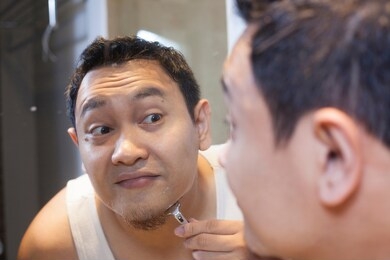 portrait of attractive young asian man shaving in bathroom, smiling and looking at mirror