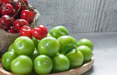 ripe fresh red cherries and green plums in bamboo bowl on white wooden background, heap of may and june fruit, summer fruits concept