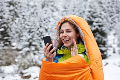 happy female waves gladfully at camera of cell phone, makes video call from top on mountains covered with snow, wrapped in sleeping bag, shares impressions about vacations, drinks hot beverage