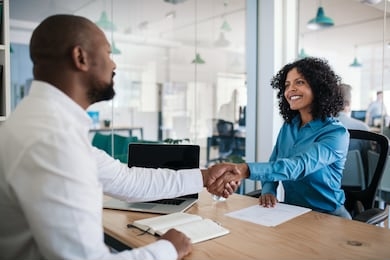 smiling african american manager sitting at his desk in an office shaking hands with a job applicant after an interview