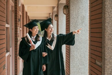 happy to be graduated. two asian female college graduates in graduation gowns walking along university corridor and talking in red brick traditional building. girl point finger showing friend view.