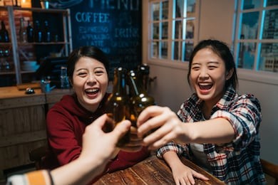 three beautiful female friends clinking bottles of beer and smiling while resting at night pub. group of happy young people cheers celebrating cheerful laughing sitting in late midnight dark bar.