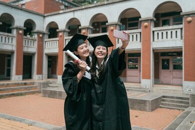 graduation day group of asian female best friends smile for self portrait looking at smart phone. two young girls cheerful showing diploma taking selfie in traditional red brick building in campus.