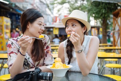 two young asian female friends travelers having fun and eating ice cream. cheerful women tourists eating mango shaved ice in summer hot outdoors using spoon sit in local cafe shop. cool fresh fruit