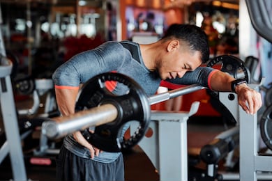 tired young vietnamese man standing at workout bench and leaning on barbell after doing exercise