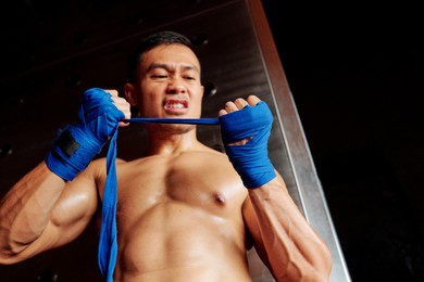 asian shirtless boxer preparing for fight and wrapping his fists wth angry expression in gym