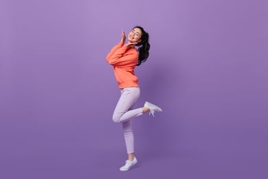 refined asian woman standing on one leg. full length view of blissful chinese girl dancing on purple background.