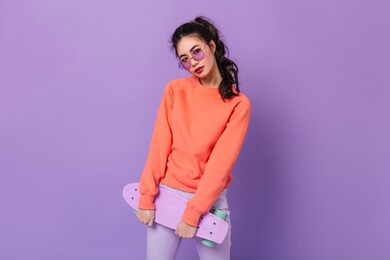 charming asian girl with ponytail holding skateboard. studio shot of adorable chinese young woman posing on purple background.