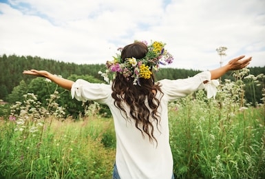 summer lifestyle portrait of beautiful young brunnete woman with perfect long curly hair in a wreath of wild flowers. standing back in the flower field, hands to the side. romantic mood. nature lover 