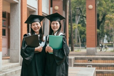 two happy asian girl college student in graduation gown and cap holding textbook learning in university finished education. women friending face camera smiling confident lovely cute joyful cheerful