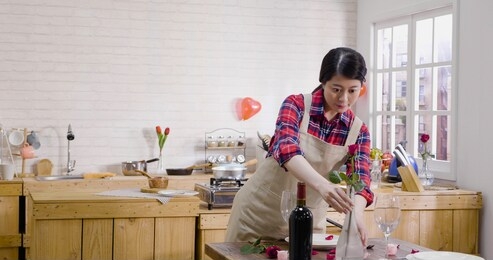 elegant young wife set up table correct arrangement of glass vase place moving flower. beautiful asian woman prepared for romantic valentine's day dinner at home indoors with wine roses glasses.