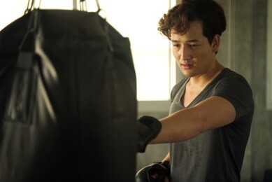 young mix-raced handsome man working out by boxing sandbag in the gym. he is punching by his left hand. seen behind the sand bag focus on the face. working out in gym and fitness concept.