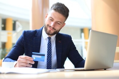 smiling man sitting in office and pays by credit card with his laptop.