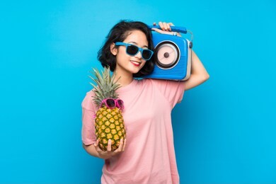 asian young woman over isolated blue background holding a pineapple with sunglasses and a radio