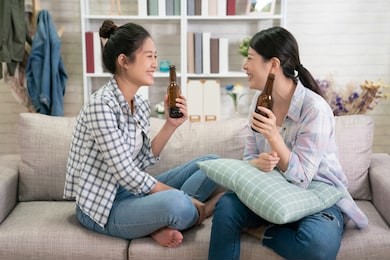 group of young asian girl friends sitting on sofa and discussing something together with beer at home. pretty ladies relax at house gossip in day time laughing cheerful. two women smiling friendship