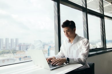 asian man sitting by the window desk using his laptop doing work