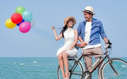 happy asian couple riding bicycle and holding colorful balloons on the beach, lifestyle concept.