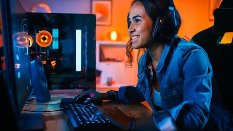 pretty and excited black gamer girl in headphones is playing first-person shooter online video game on her computer. room and pc have colorful neon led lights. cozy evening at home.