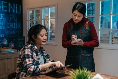 asian waitress takes order in restaurant using note and pen writing standing talking with client in night pub in modern city. cute girl customer point on digital tablet screen choose meal in bar.