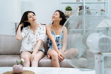 smiling young girl roommates with air cooler relax in comfort temperature at home. lazy family weekends sisters enjoy electric fan sitting on couch in living room on summer day hot weather outdoors