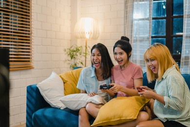 group of asian women couple play games at home, female using joystick having funny moment together on sofa in living room in night. teenager young friend football fan, celebrate holiday concept.
