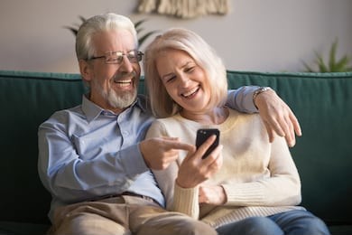 happy senior old couple holding smartphone looking at cellphone screen laughing relaxing sit on sofa together, smiling elder mature grandparents family embracing having fun with mobile phone at home
