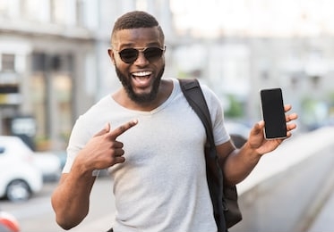 hey something new. happy african guy pointing his cellphone with black blank screen for advertisement outdoor, panorama