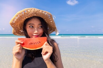 beautiful young asian woman holding watermelon, looks at copy space in summer season at beautiful beach in hot weather that make beautiful asia lady eat watermelon to cool down, quench her thirst
