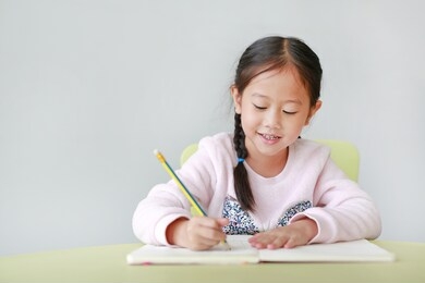 happy little asian child girl writes in a book or notebook with pencil on table in classroom against white background.