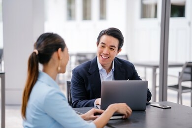 a young, handsome and well-groomed asian man in a dark suit is interviewing a professional woman candidate applying a job during the day. he is smiling as he talks to the woman. 