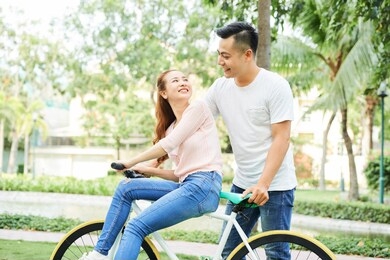asian smiling woman sitting on bicycle and looking at her boyfriend while he riding her in the park
