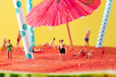 some miniature people wearing swimsuit relaxing on a refreshing ripe watermelon, with some white and blue drinking straws and a paper umbrella stuck in it, against a yellow background