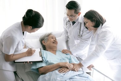 elderly female smiling with two young doctor and young female nurse writing in clipboards visiting asian senior patient woman at hospital ward - senior medical and healthcare concept