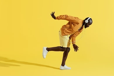 man in headphones listening music, dancing on yellow background. full length portrait of handsome black male model in colorful fashion clothes and white earphones in studio