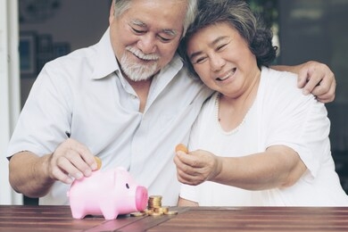cheerful elderly couple saving money with putting coin into piggy bank and stack of coin on wooden table, they are smiling with happiness together