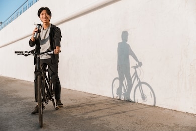 smiling asian man riding a bicycle outdoors, drinking water from a bottle