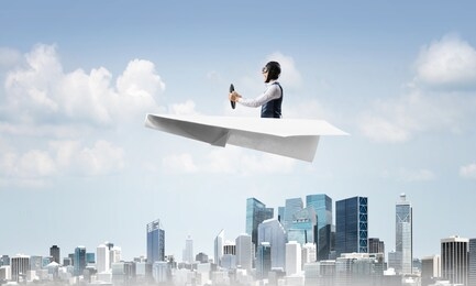 aviator driving paper plane above business center in cloudy blue sky. pilot in leather helmet sitting in paper plane and holding steering wheel. cityscape with high skyscrapers and office buildings.