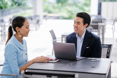 a chinese asian manager in a suit has a meeting with his colleague, a woman in a pale blue suit. he is conducting a performance appraisal during this meeting. they are smiling and talking. 
