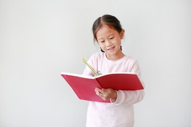 smiling little asian child girl writes in a book or notebook with pencil in classroom against white background.